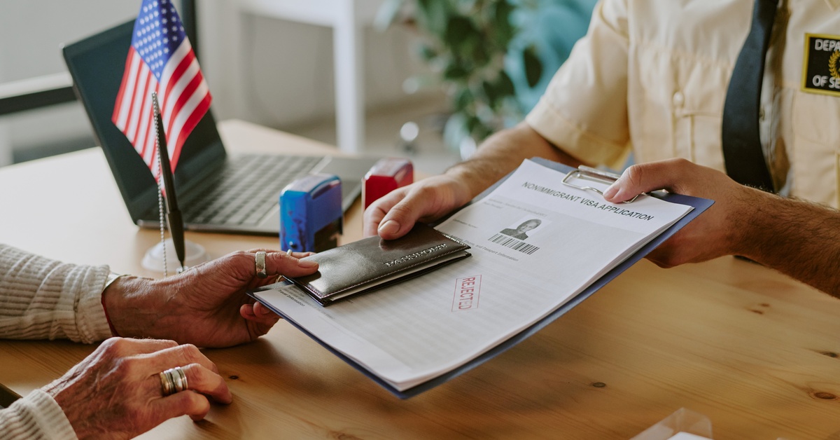 A person wearing a brown security uniform hands over documents and a passport to another person across the table.