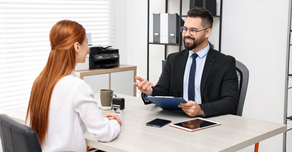 A man wearing a black suit smiles while holding a blue clipboard and sitting across from a woman wearing a white cardigan.