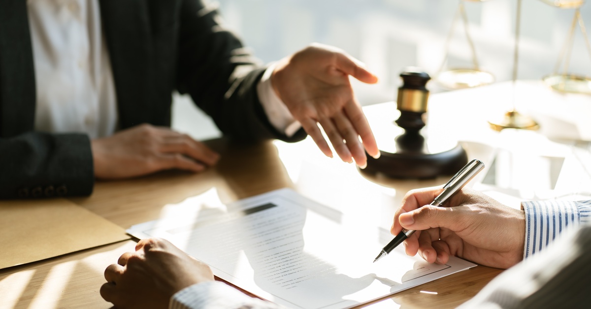 A person writing on a document with a silver pen, sitting across from another person, gesturing toward the document.