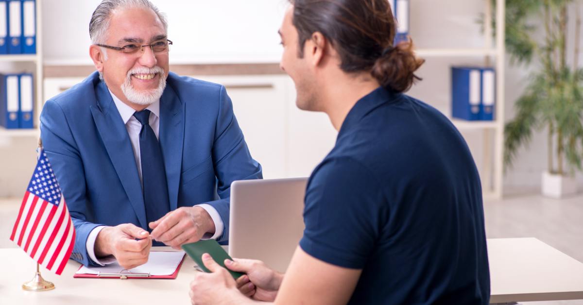 A person wearing a blue shirt smiles while sitting across another person wearing a suit. On the table is the American flag.
