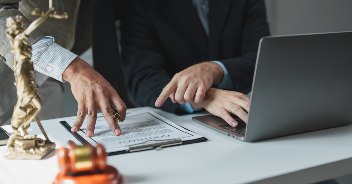 A man is typing on a laptop while another man next to him points to a contract on a clipboard.