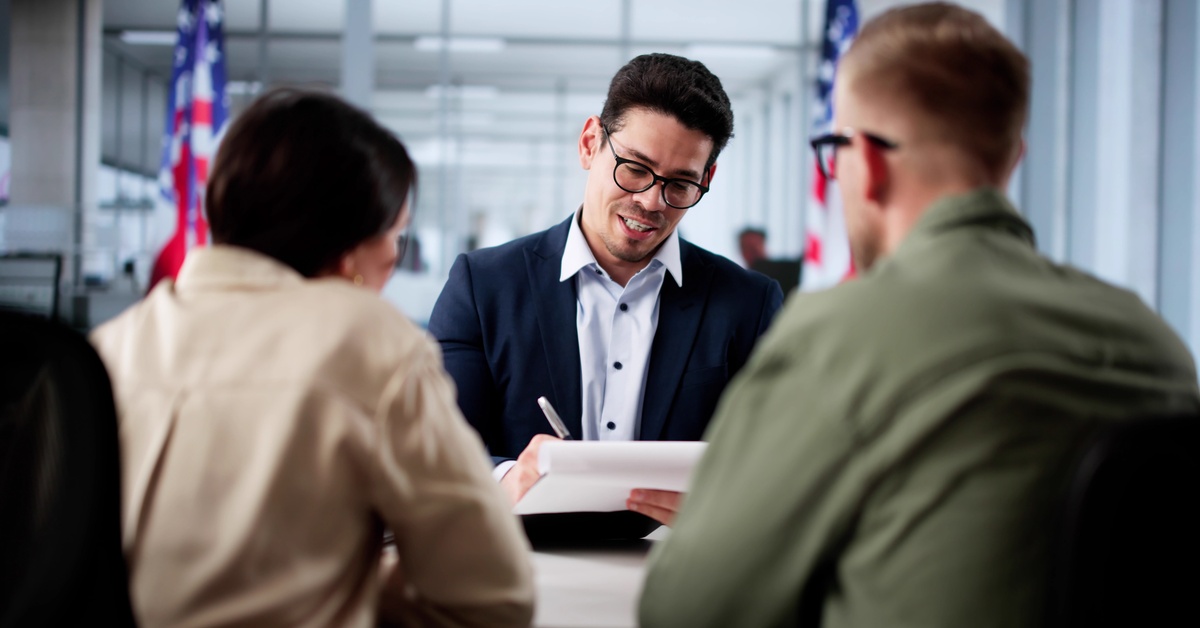 Two people sit across a man with eyeglasses, wearing a blue suit, while writing on a document with a pen.