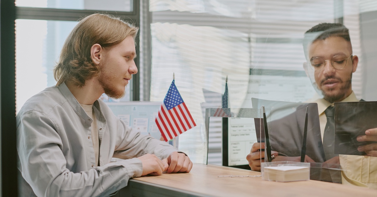 A man is interviewing another man while holding a passport holder in a booth with an American flag next to it.