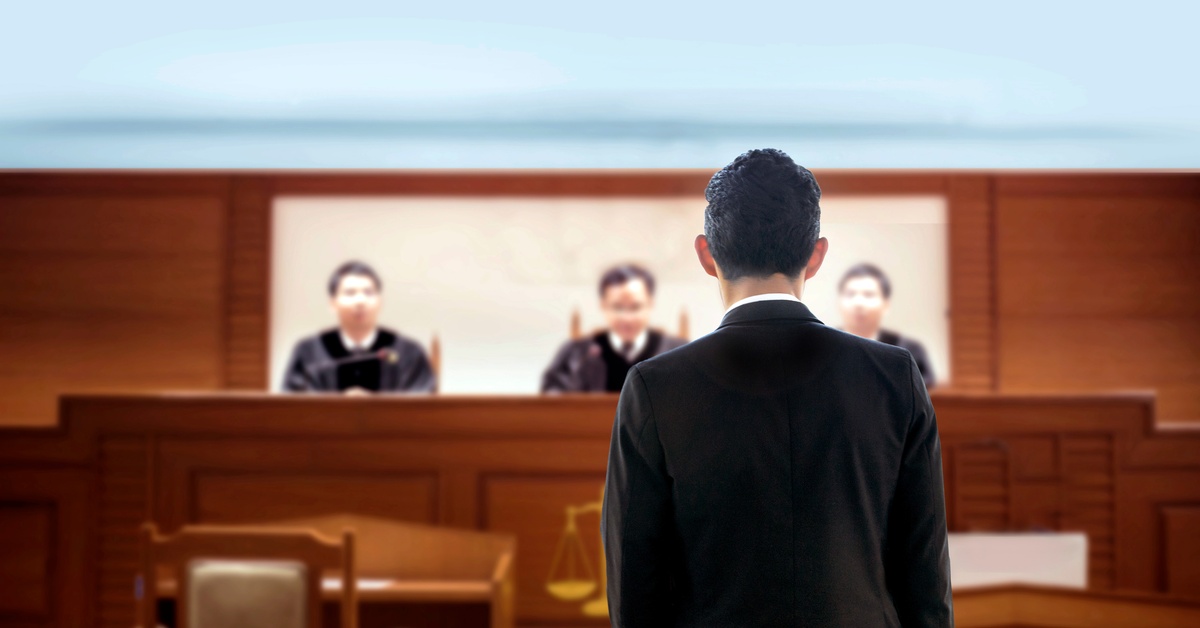 A man in black suit stands with their back turned in a courtroom. In front of him are three magistrates.