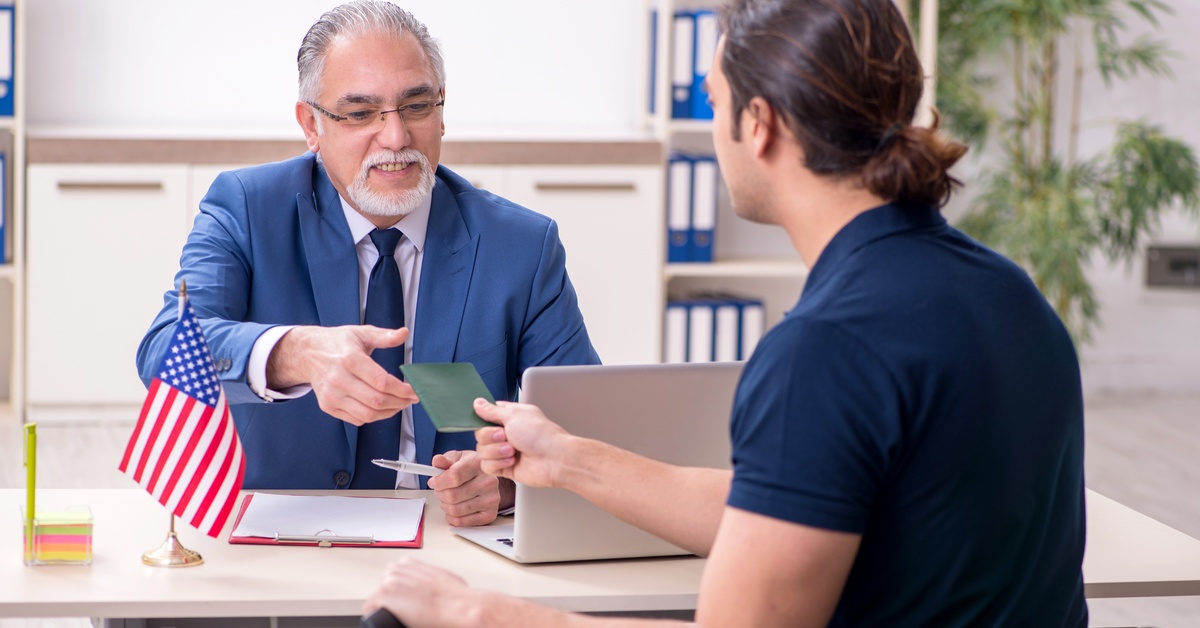 A man holding a suitcase gives a green passport book to an older man wearing a suit sitting across him.