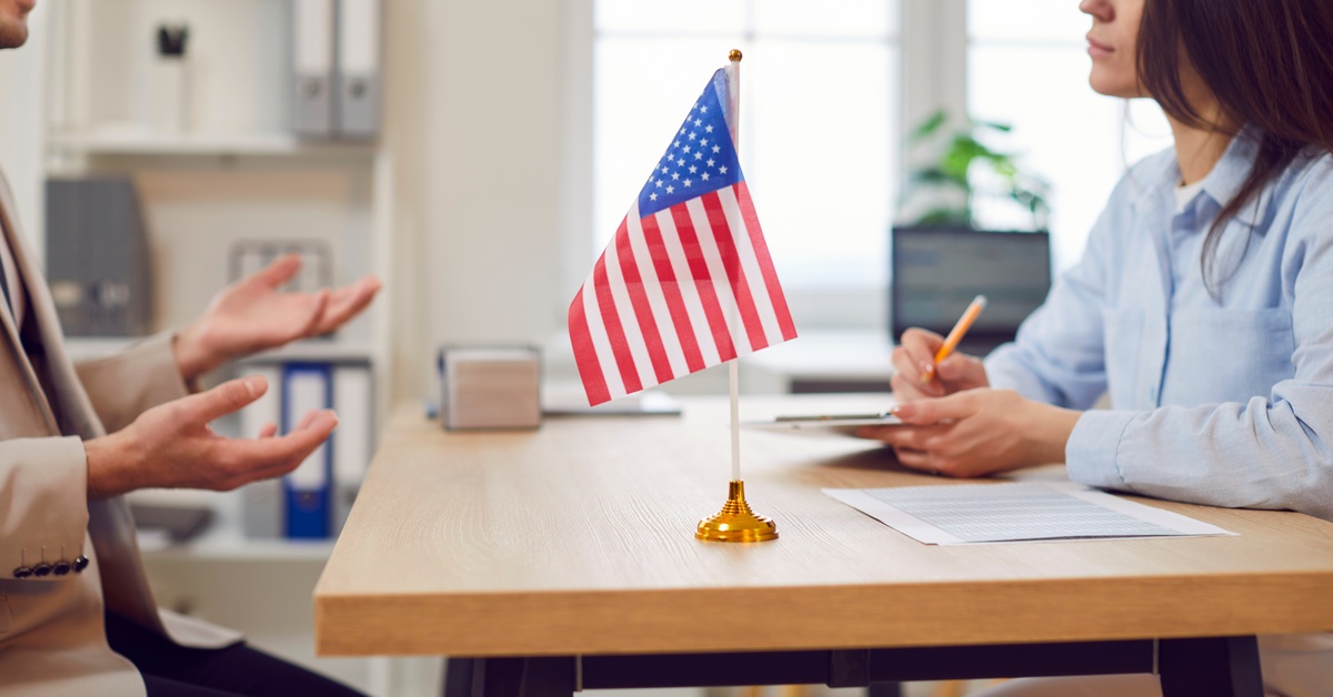 A woman holding a clipboard and a pencil listens to another man sitting across the table with the American Flag.