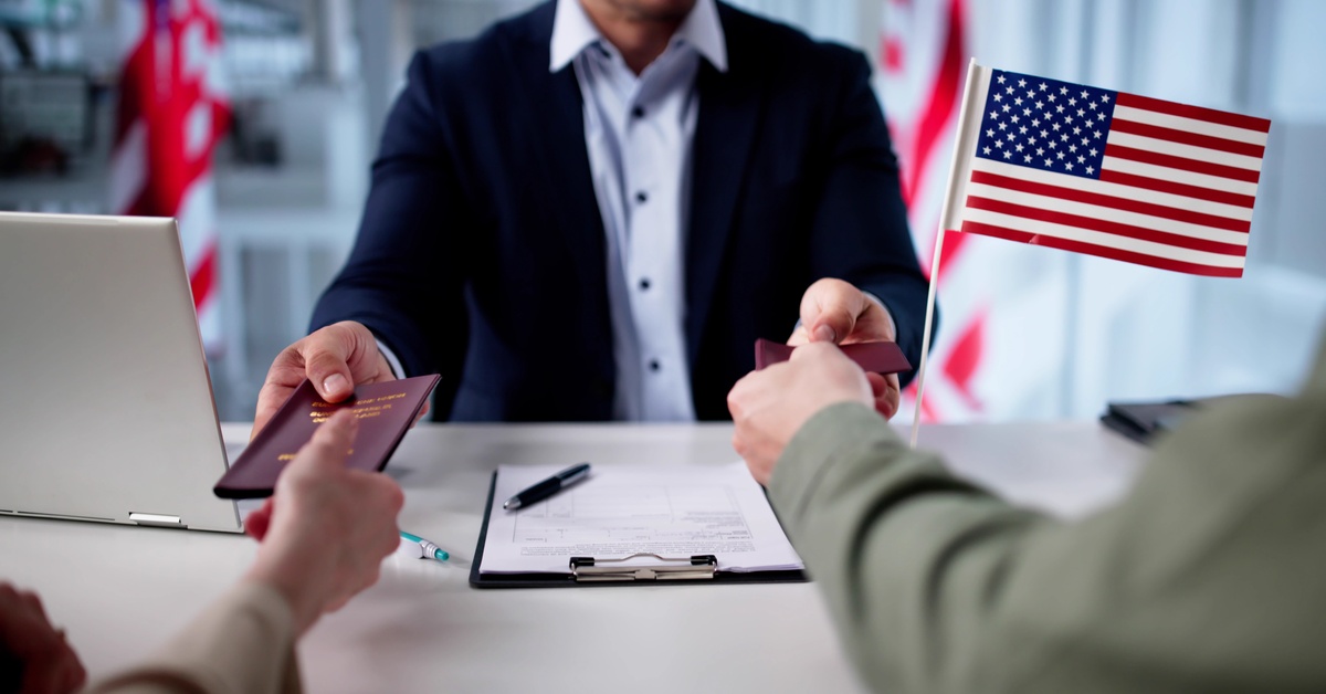 An attorney hands over passports to the two people sitting in front of them, next to an American flag.