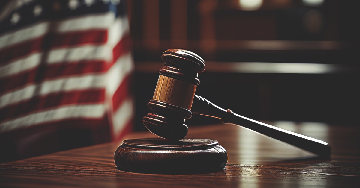 The light shines down on the edges of a wooden gavel sitting on top of a table, with the American flag behind it.