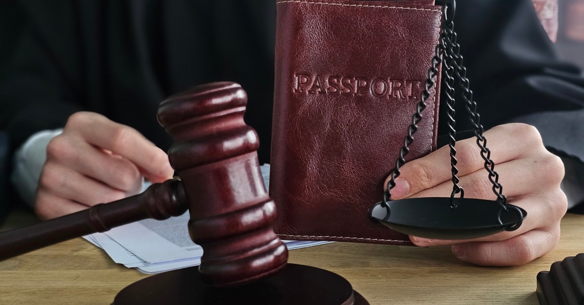 A judge holds a leather passport next to a wooden gavel and a black scales of justice placed on the table.