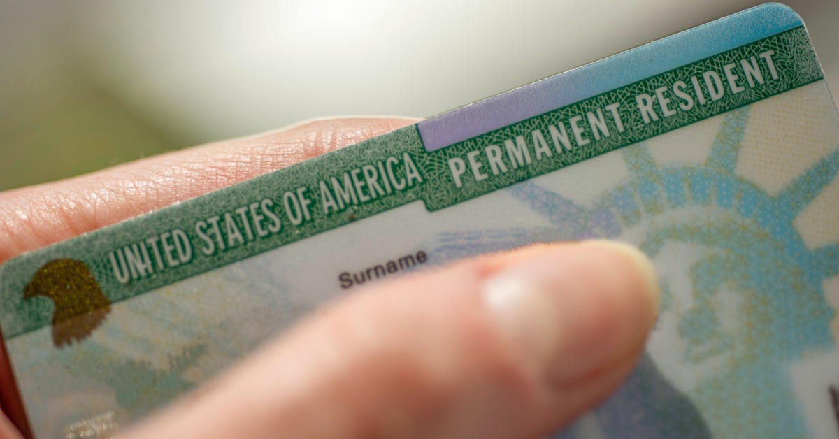a close-up view of a hand holding a green permanent resident card in front of a blurred background behind it.