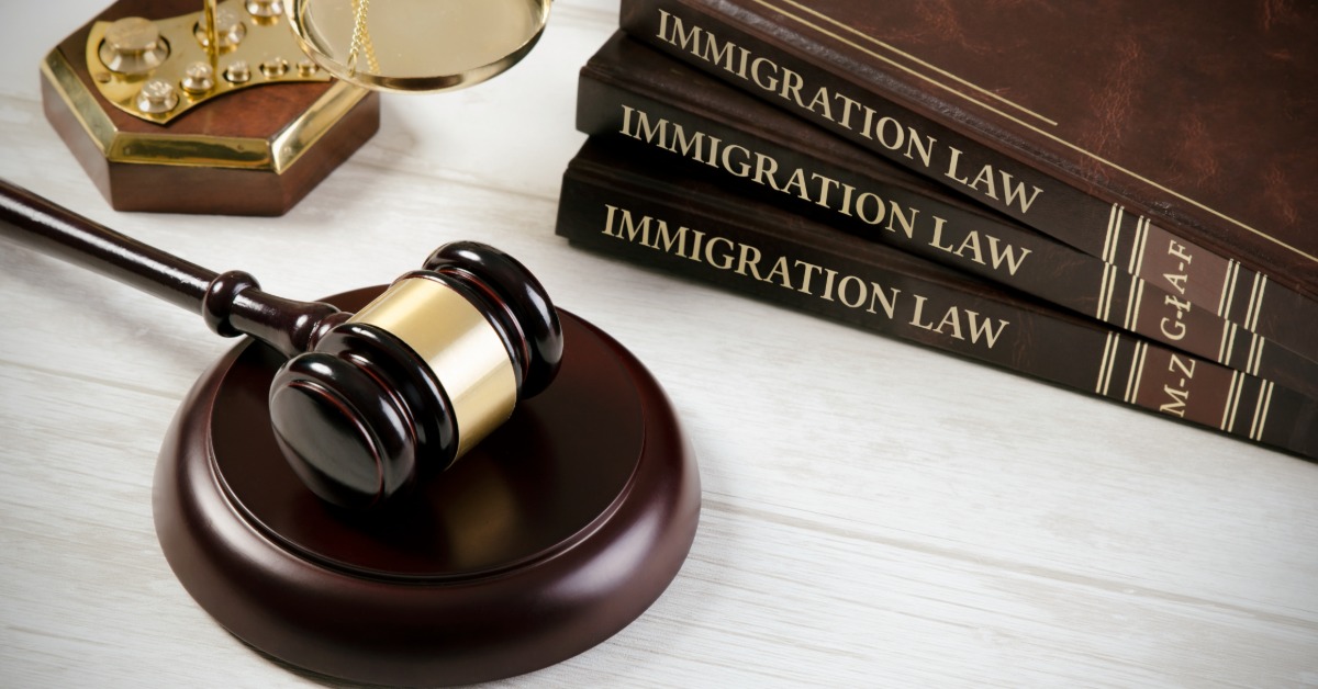 a wooden gavel rests near a balance scale and a stack of books labeled Immigration Law on a light desk.