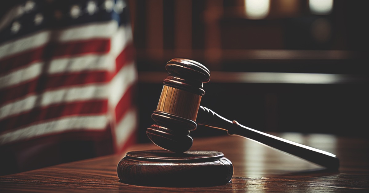 a wooden gavel rests on a block across a polished desk, with a blurred American flag in the background.