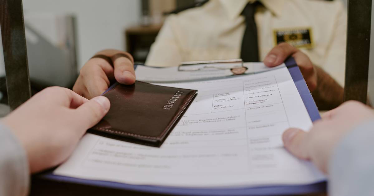 Hands passing a passport and paperwork across a service counter toward a uniformed person behind glass.