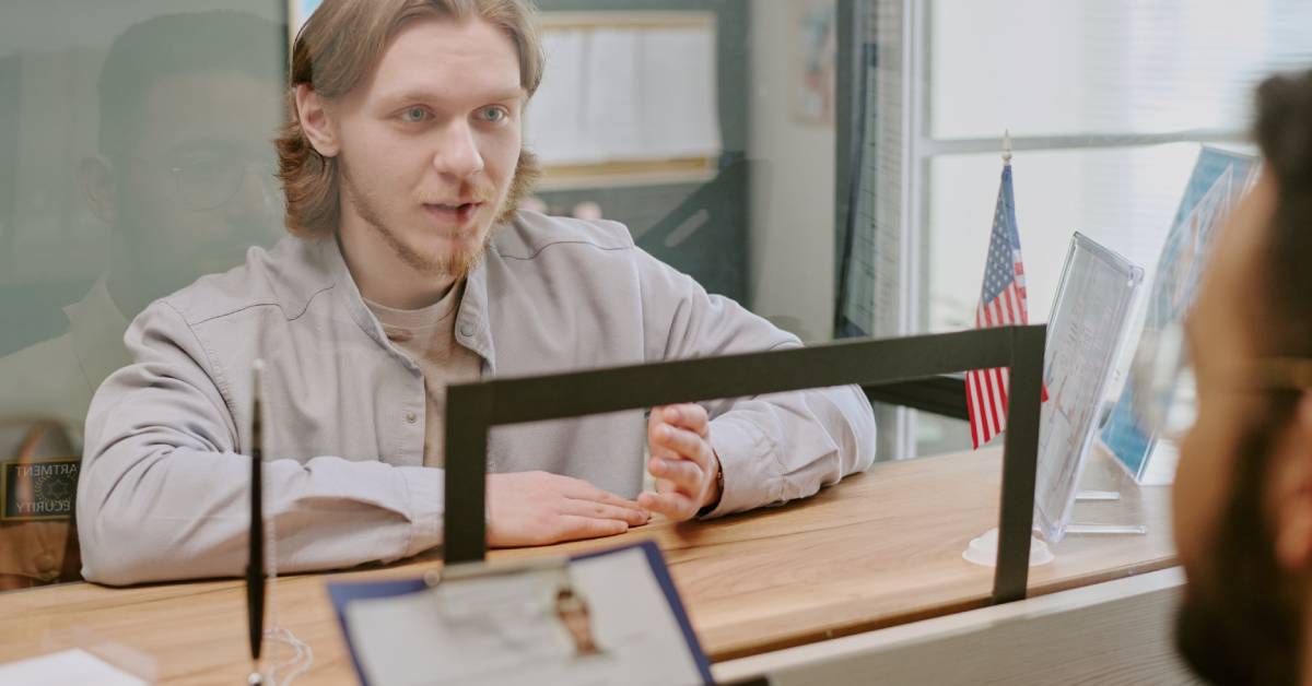 A man seated behind glass at a service counter with a blurred person in front and a small American flag nearby.