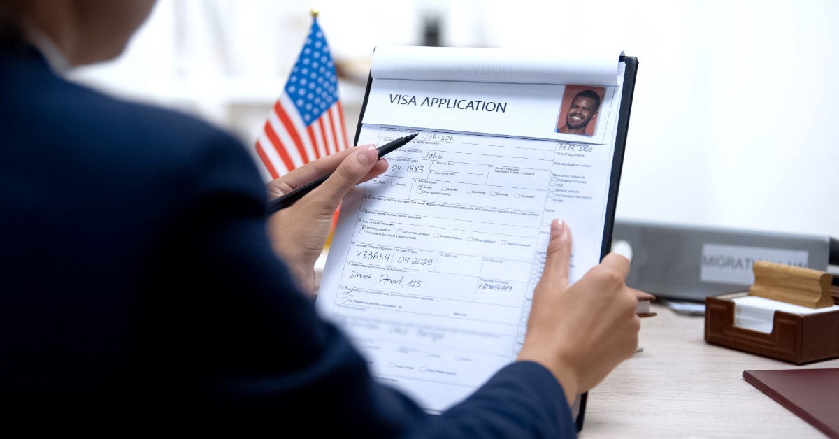 A person holding a pen and visa application on a clipboard, with a small American flag and desk items nearby.