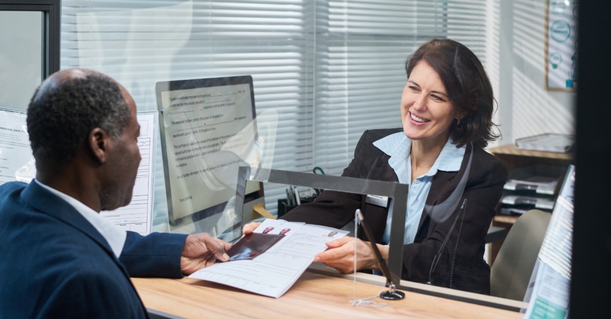 A smiling woman passing documents through a glass partition to a seated man at a visa processing counter.