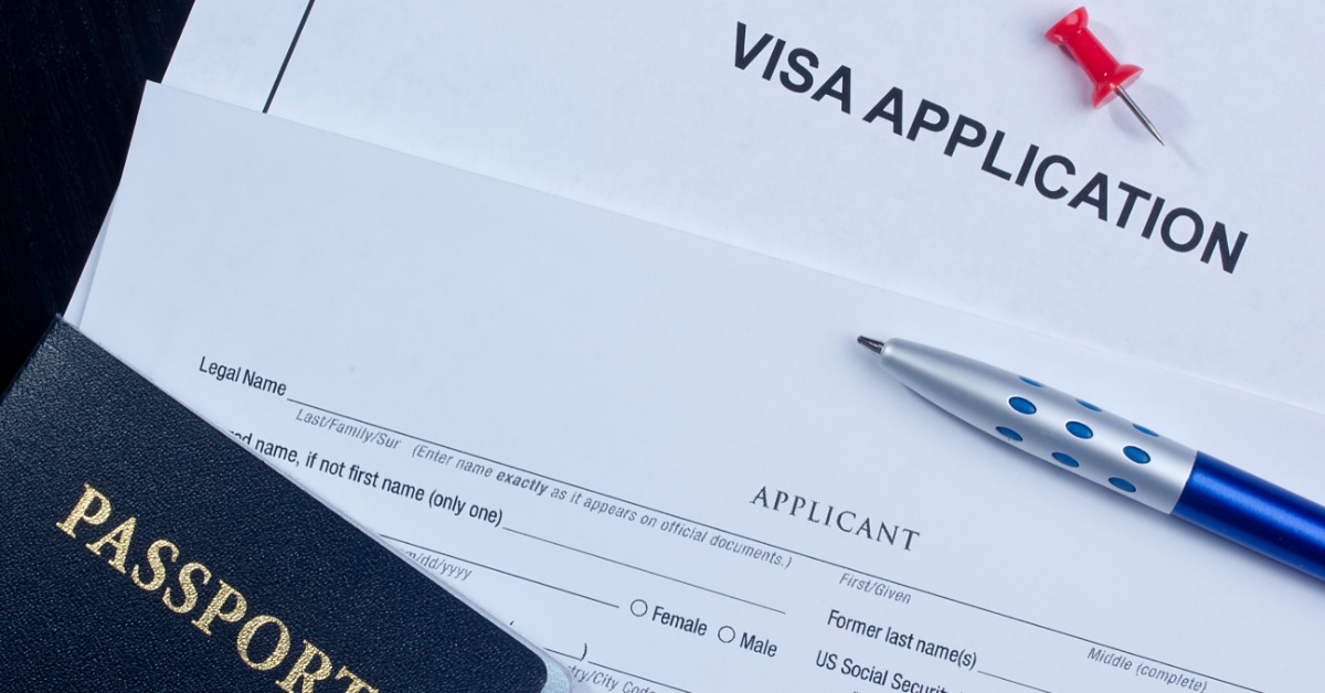 A passport, visa application paper, blue pen, and red pushpin arranged on a dark surface with blank form fields.