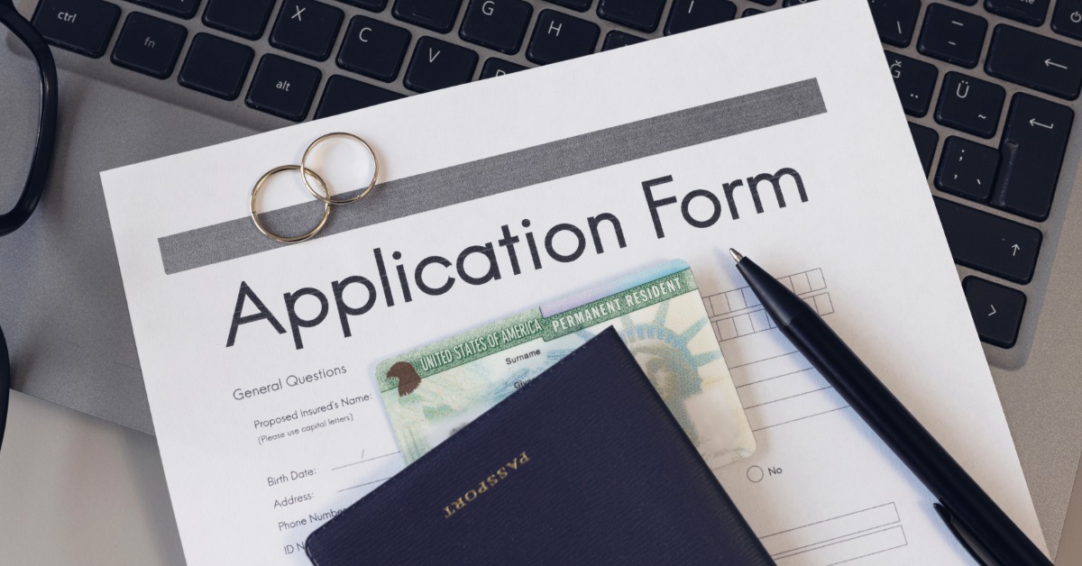a passport and green card resting on a printed application form with two rings and a pen over a laptop keyboard.