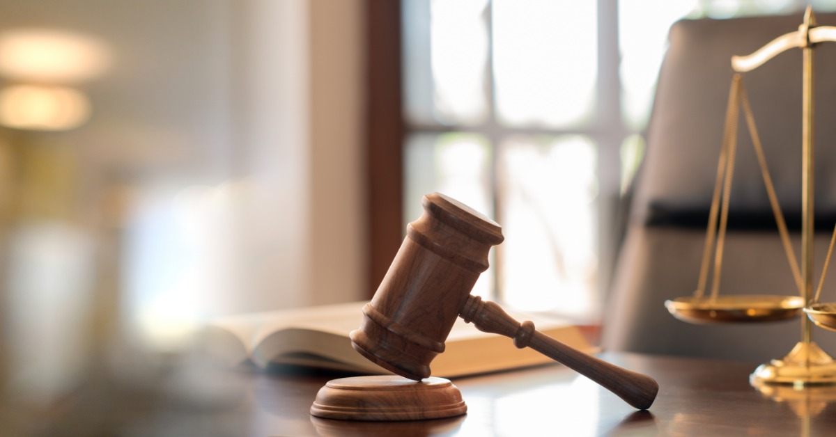 A wooden gavel and brass scales on a desk beside an open book, with chairs and windows in the background.