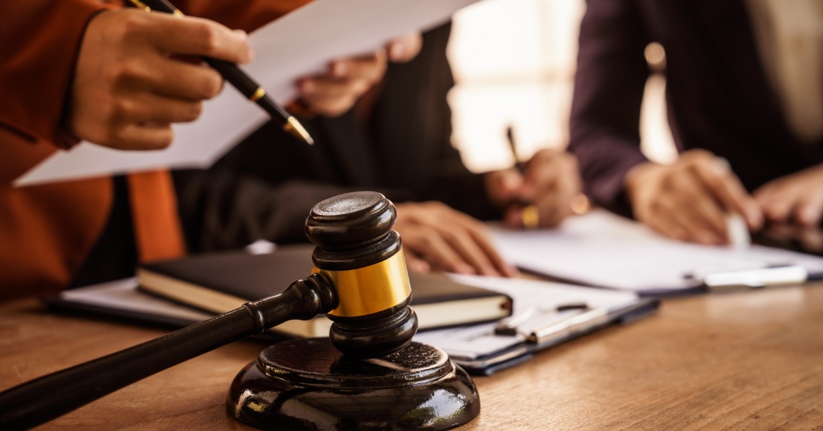 A gavel on a wooden table near people holding papers and writing on documents with pens during a meeting.